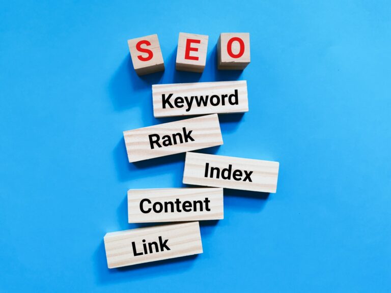 Wooden blocks spelling out SEO, Keyword, Rank, Index, Content, and Link are arranged on a blue background. The SEO blocks are on top and featured prominently in red letters.