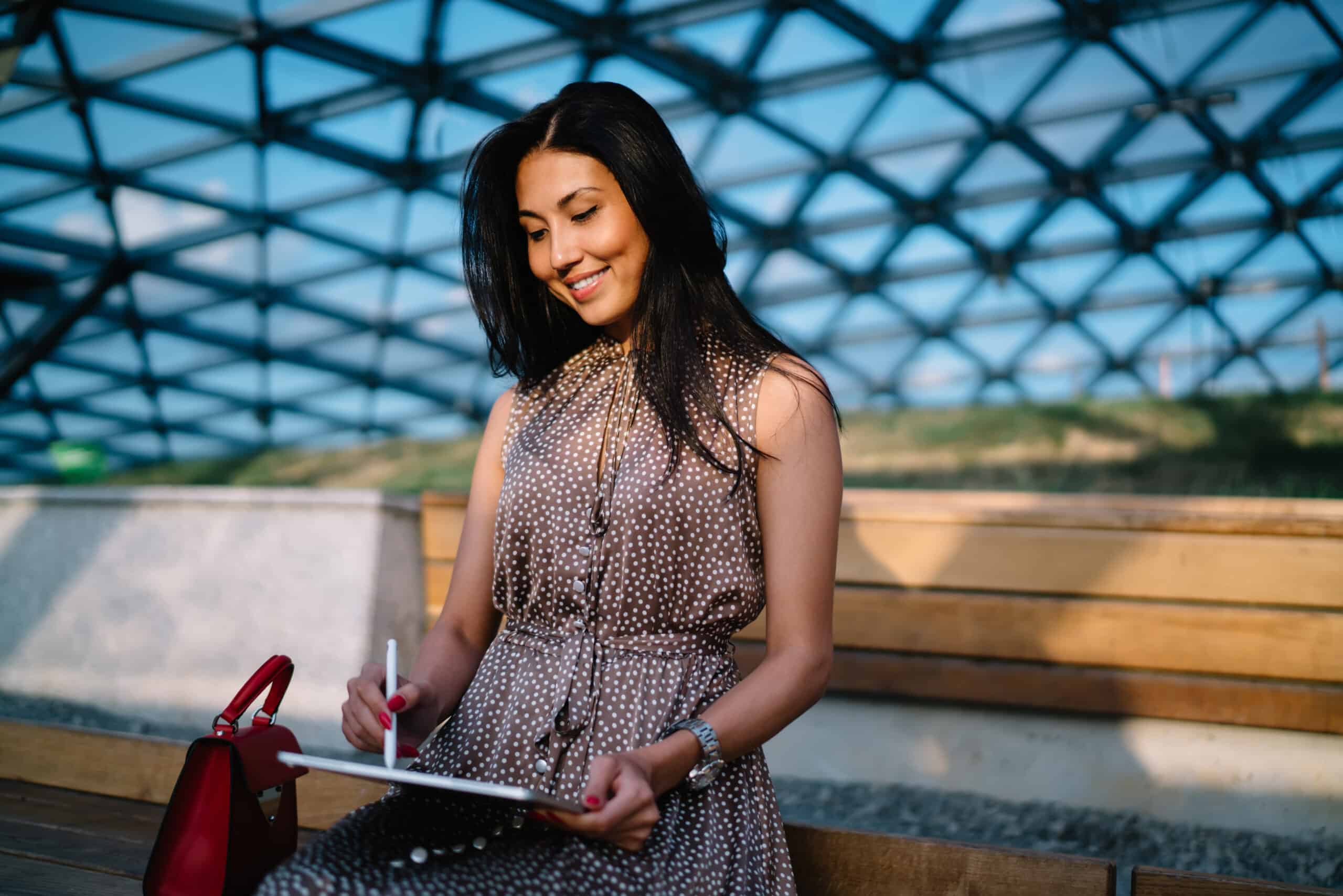 A woman in a brown polka-dot dress sits on a wooden bench, smiling while using a stylus on a tablet—perhaps building an Irish brand—outdoors under a geometric glass roof with bright natural light.
