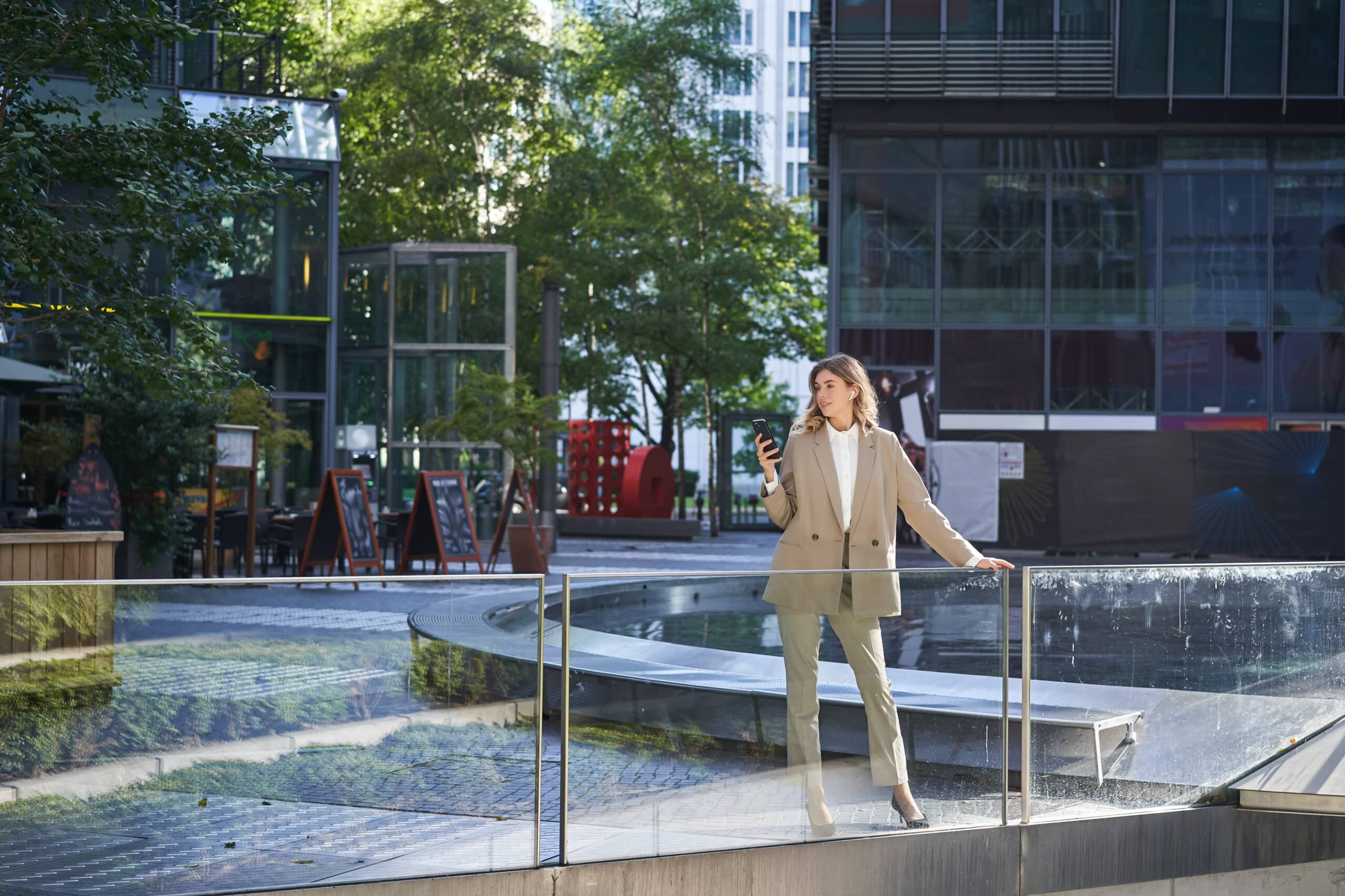 A woman in a beige suit stands outdoors near a glass railing, looking at her phone—perhaps exploring tips for building an Irish brand—with modern office buildings and trees in the background as sunlight filters through the urban plaza.
