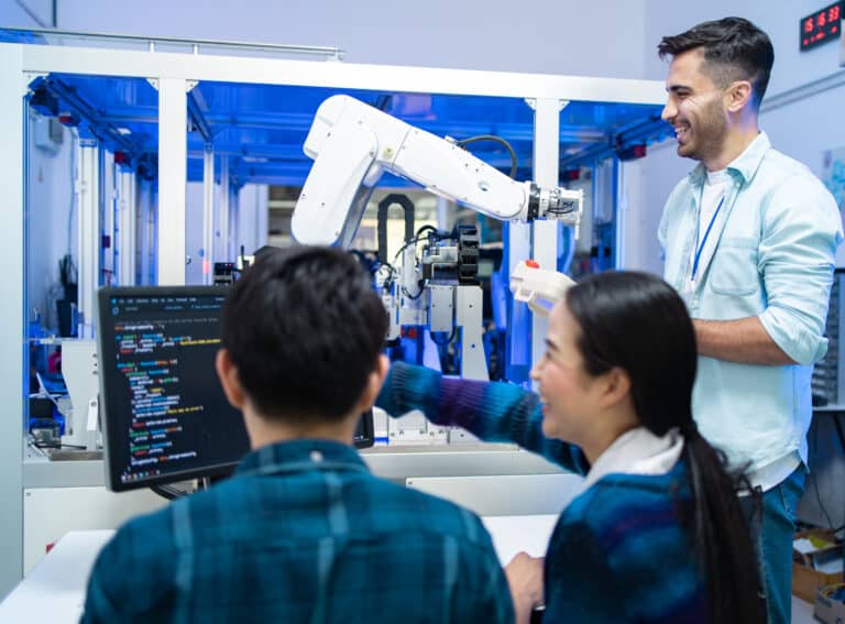 Three people collaborate in a lab with a robotic arm, building an Irish brand. Two are seated at a desk with code on the screen; the third stands, smiling, holding a device. Lab equipment and a blue-lit machine fill the background.