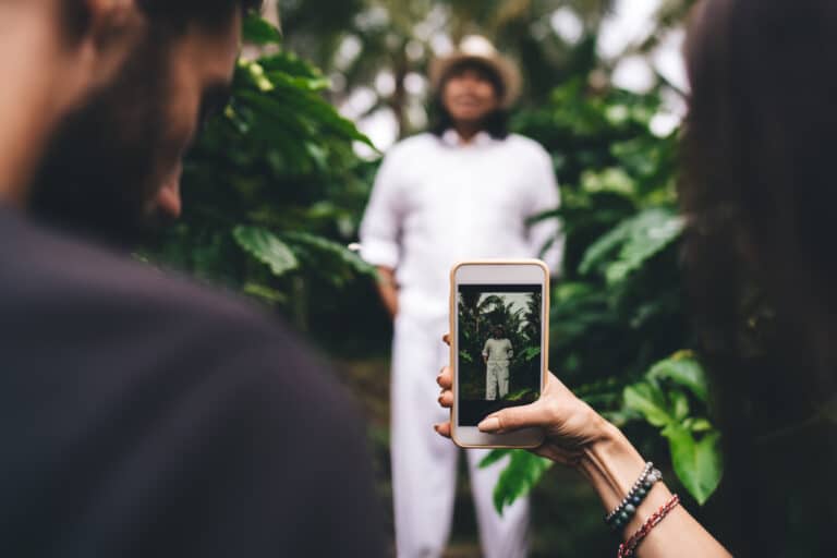 A person holds a smartphone, taking a photo of a man in white clothes and a hat standing among green plants—capturing the essence of building an Irish brand, as his image is displayed clearly on the phone screen.