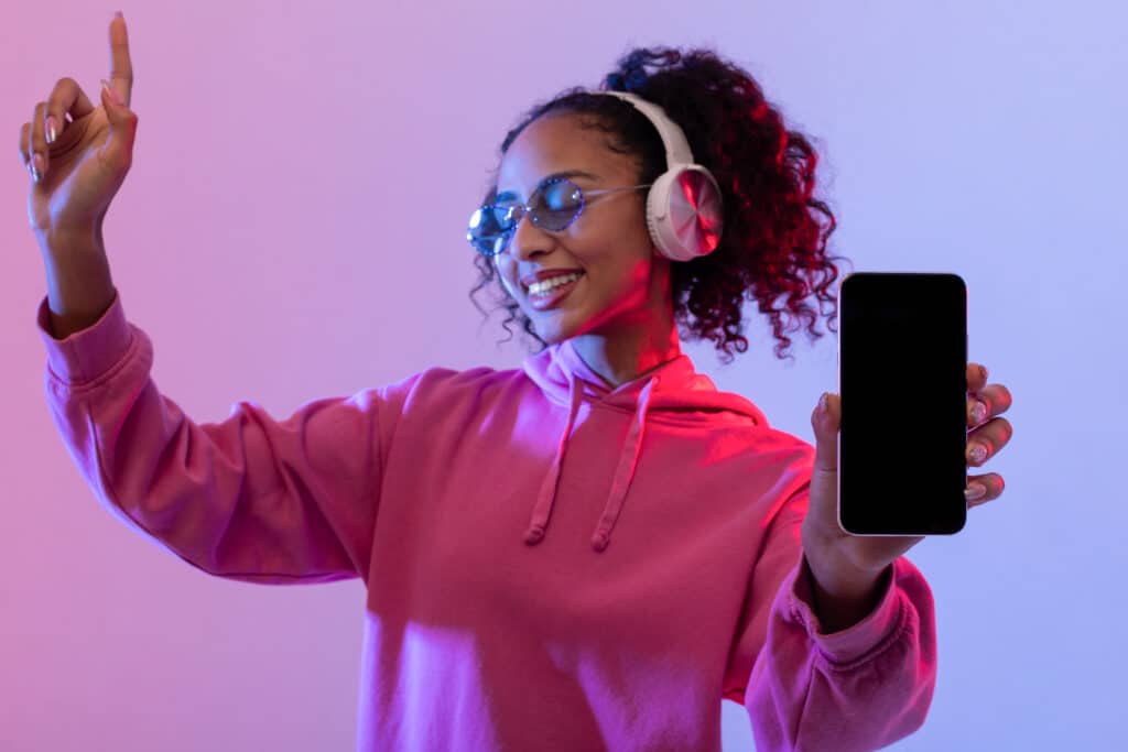 Smiling young woman wearing headphones, sunglasses, and a pink hoodie dances with one hand up while showing a blank smartphone screen against a purple and blue gradient background, capturing the energy of building an Irish brand.