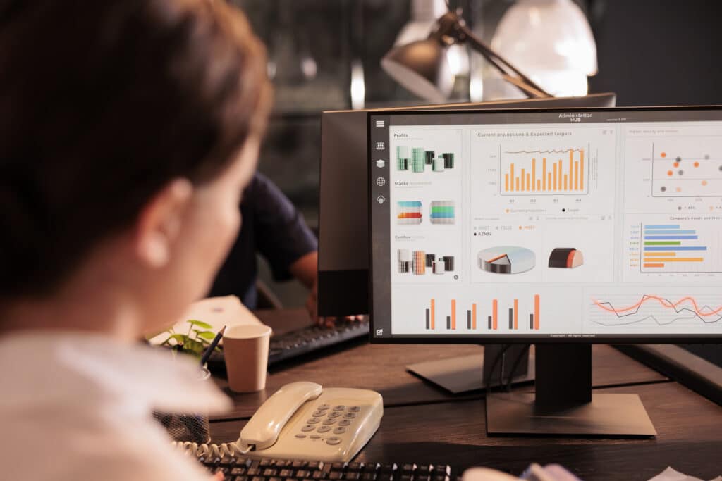 A person at a desk looks at a computer monitor displaying colorful charts, graphs, and data visualizations while building an Irish brand in an office setting. A telephone, keyboard, and coffee cup are also on the desk.