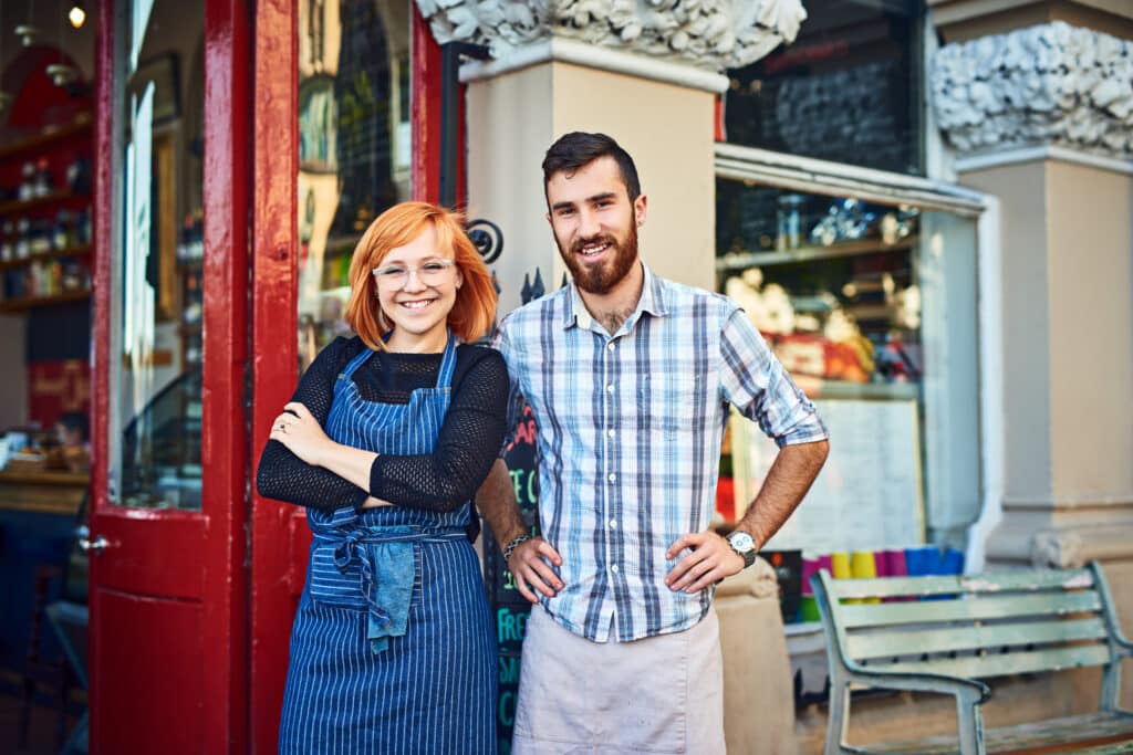 Two young adults, one in an apron and glasses and the other in a plaid shirt, stand smiling outside a shop with a red door—capturing the spirit of building an Irish brand in their inviting café.
