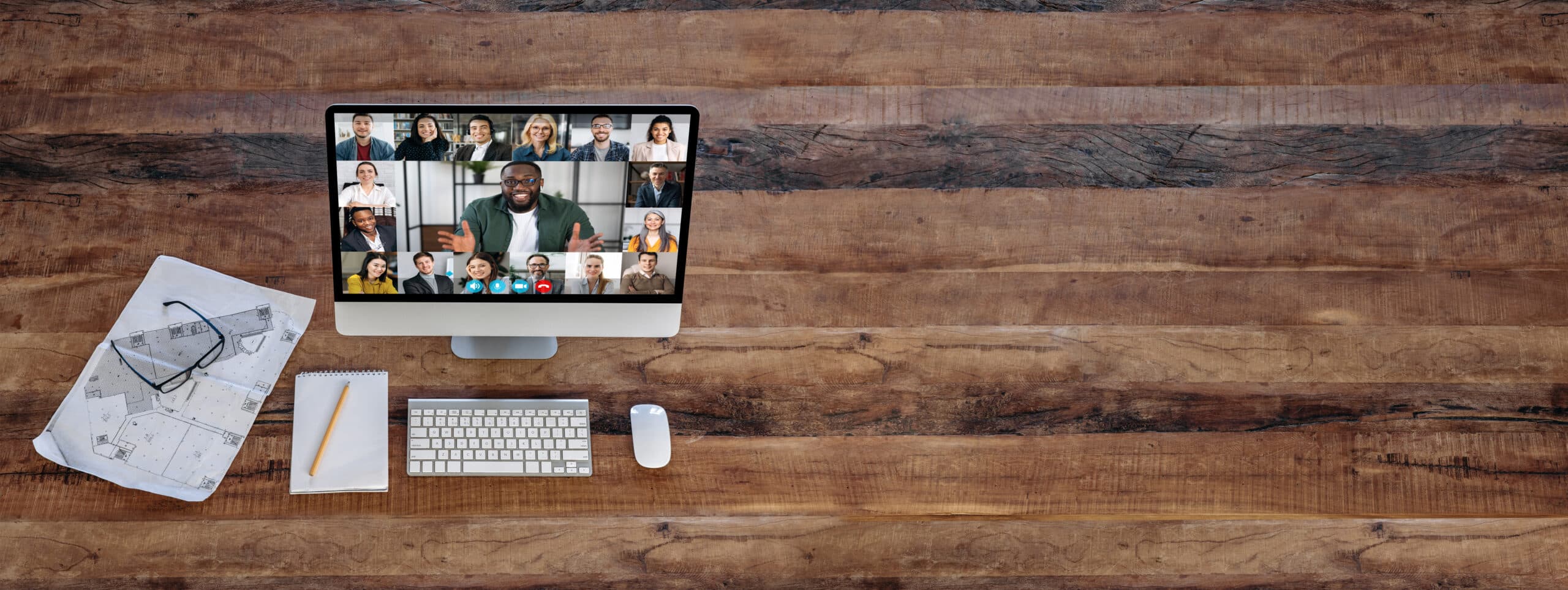 A computer on a wooden desk displays a video call with many people. Nearby are architectural plans, glasses, and a pencil—tools for building an Irish brand. The rustic desk has plenty of empty space, ideal for creative work.