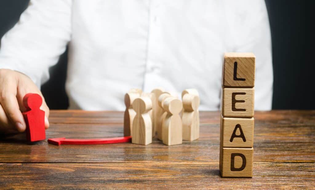 A person in a white shirt moves a red wooden figure toward beige figures, with blocks stacked to spell LEAD on a wooden table, symbolizing leadership and guidance in building an Irish brand.