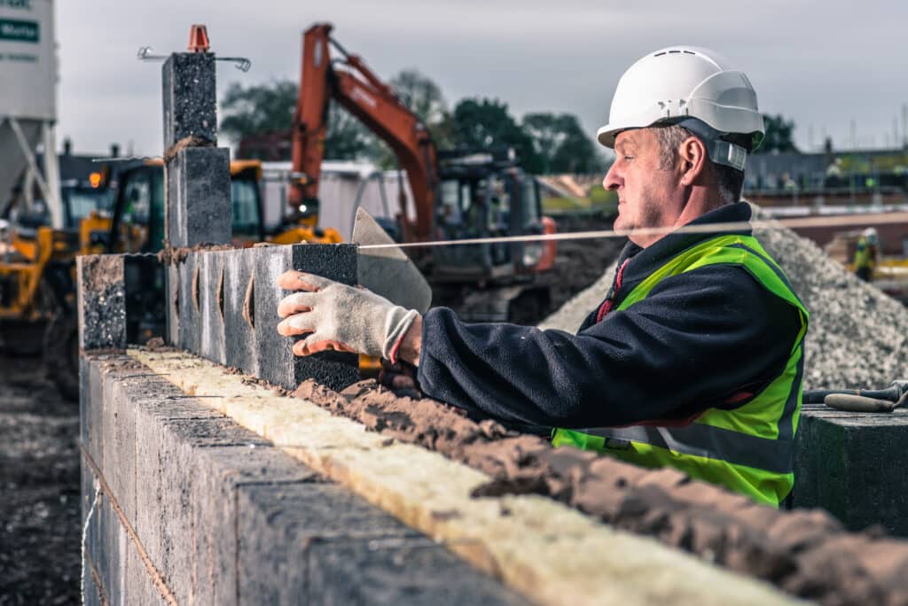 A construction worker wearing a white hard hat and high-visibility vest carefully places a concrete block onto a wall at a construction site, building an Irish brand with machinery and building materials in the background.