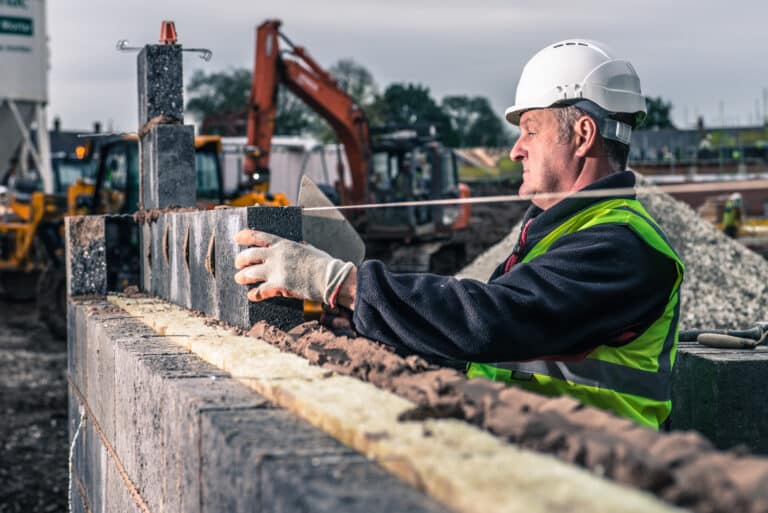 A construction worker wearing a white hard hat and high-visibility vest carefully places a concrete block onto a wall at a construction site, building an Irish brand with machinery and building materials in the background.