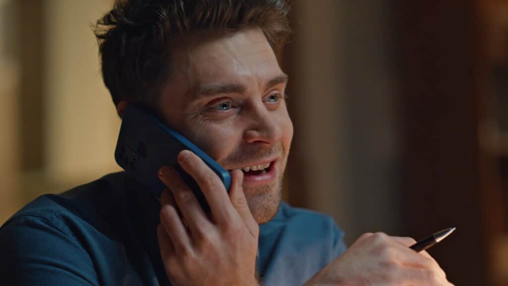 A smiling man with light brown hair holds a phone to his ear and a pen in his other hand, appearing engaged in a cheerful conversation about building an Irish brand. He wears a blue shirt and is indoors, with a warm, softly lit background.