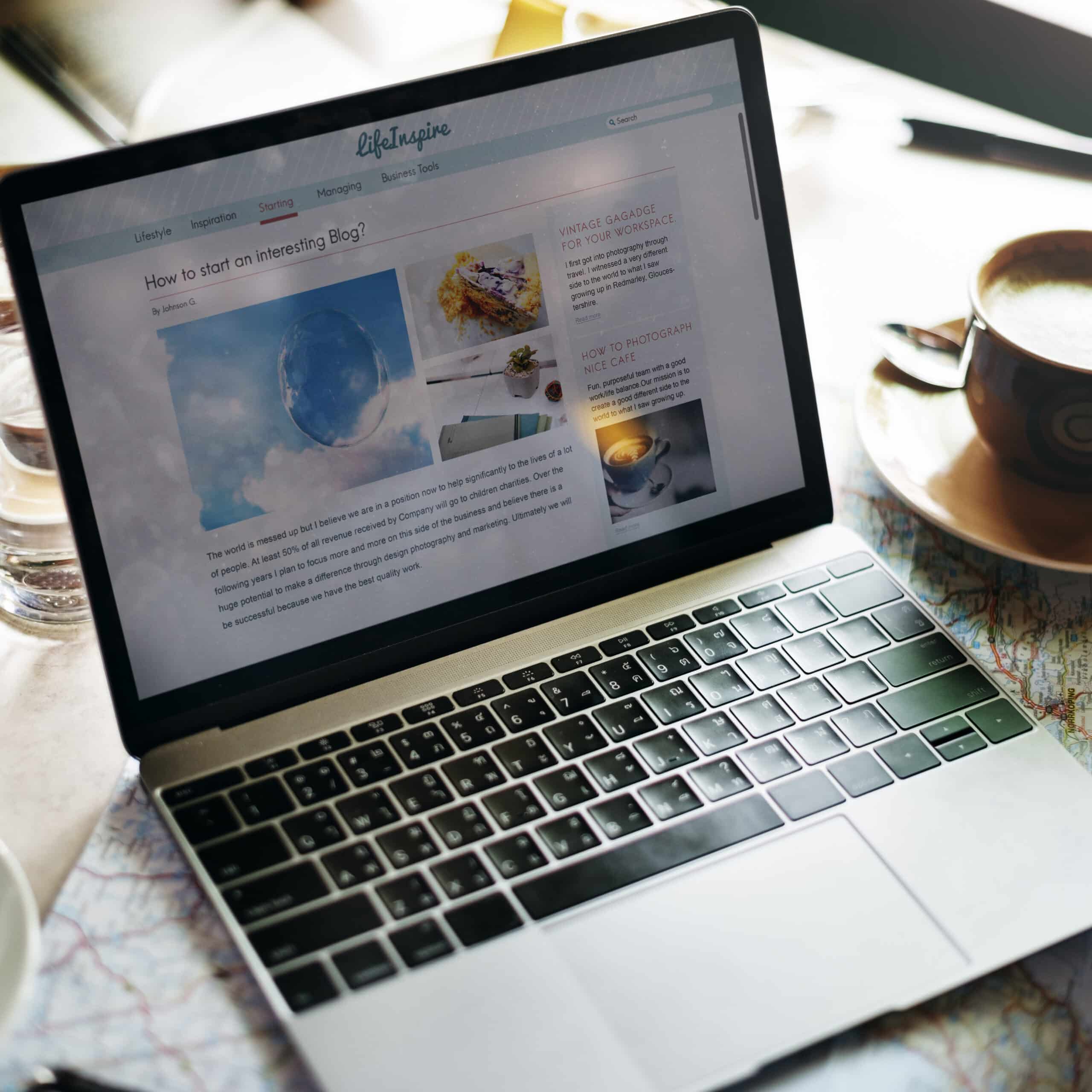 A laptop on a table displays a blog article titled “How to start an interesting Blog?” next to a cup of coffee, a saucer, a glass of water, and a map - perfect inspiration for building an Irish brand.