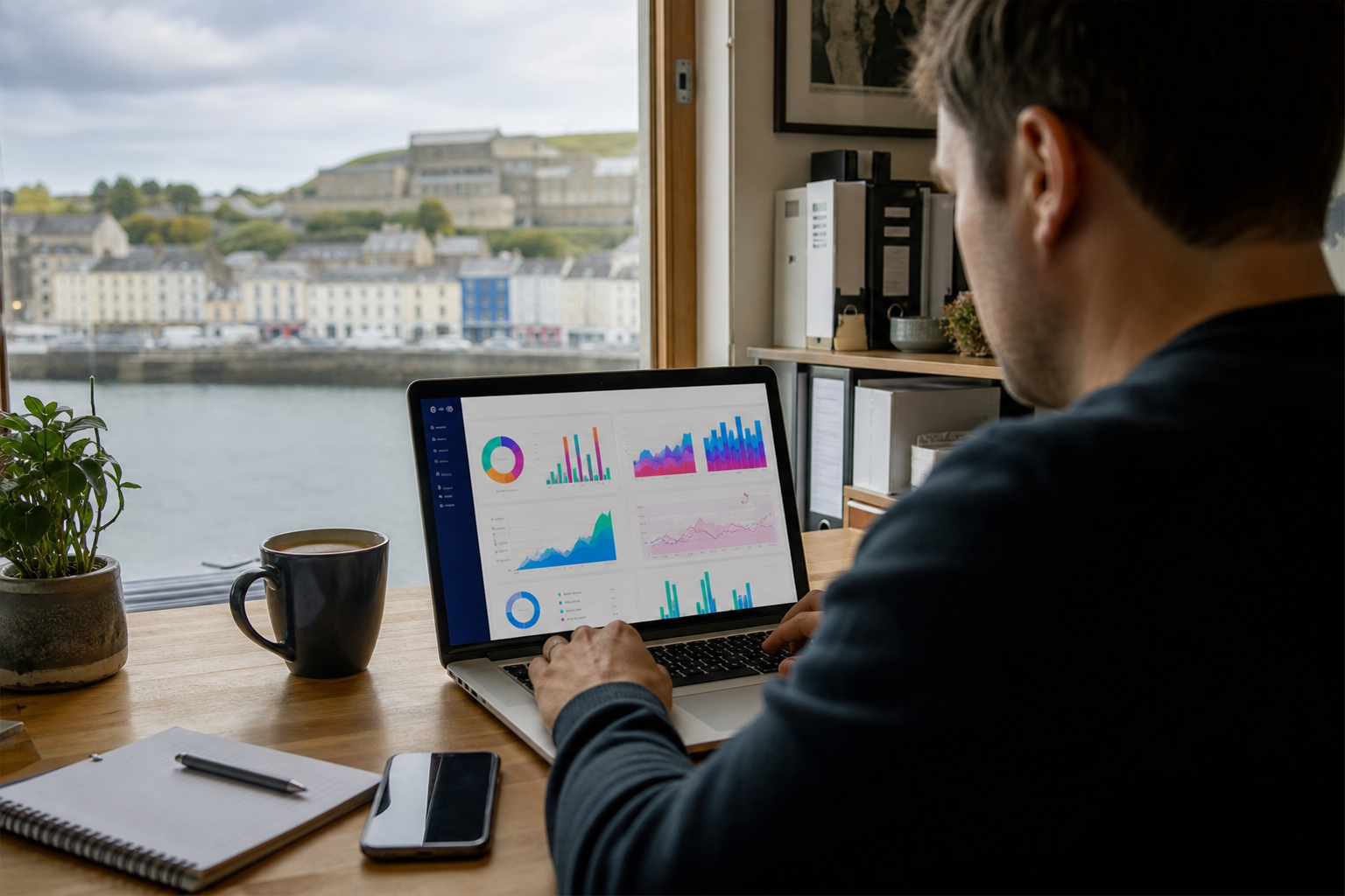 A person sits at a wooden desk working on a laptop displaying various data charts, with a view of a waterfront town visible through the window.