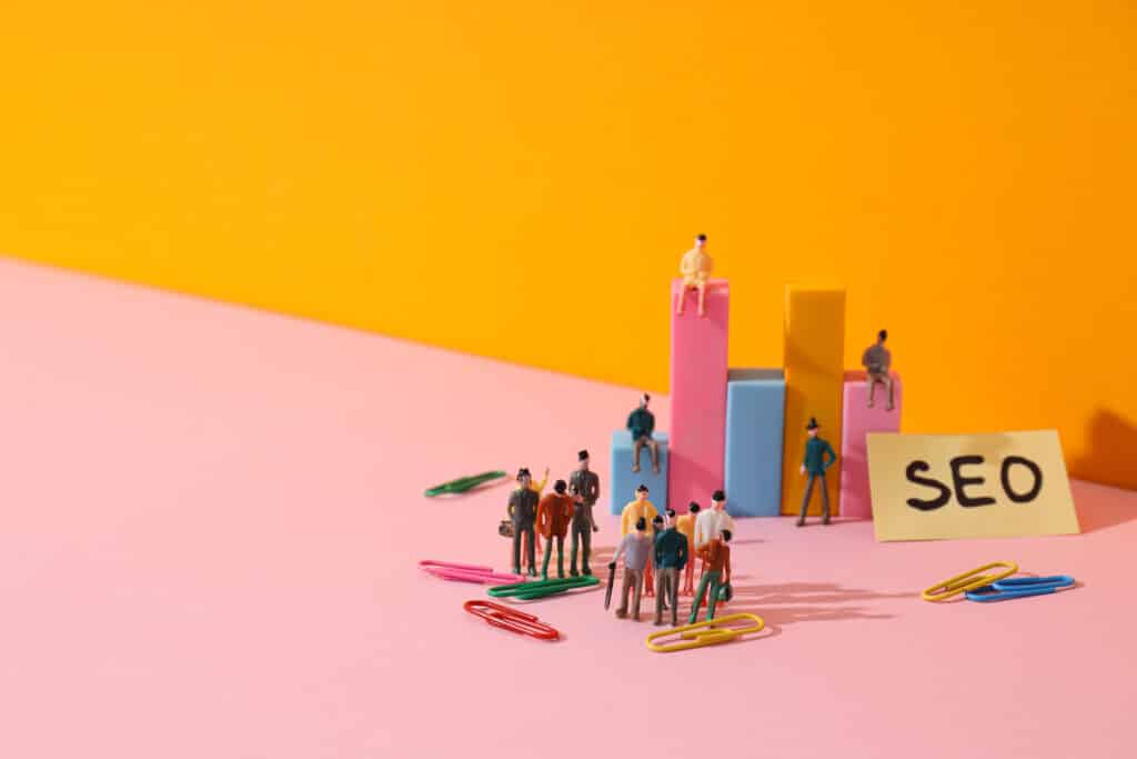 Miniature figures stand near colorful paperclips and bar graphs on a pink surface with an orange background. A yellow sticky note labeled SEO sits beside them, symbolizing building an Irish brand through SEO and digital marketing concepts.
