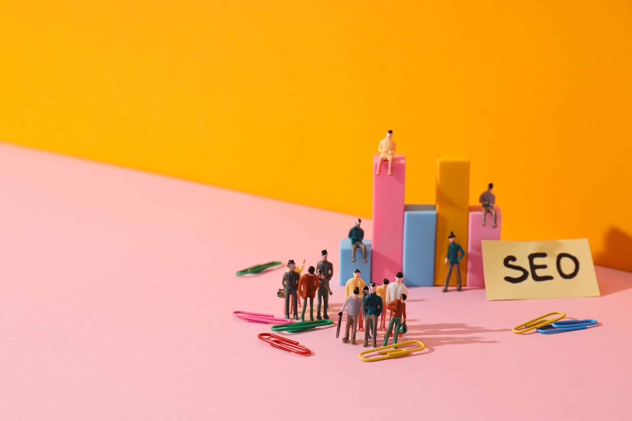 Miniature figures stand near colorful paperclips and bar graphs on a pink surface with an orange background. A yellow sticky note labeled SEO sits beside them, symbolizing building an Irish brand through SEO and digital marketing concepts.